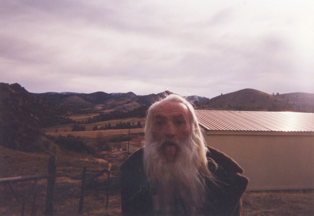 An older man with wild white hair and beard sticks his tongue out to the camera