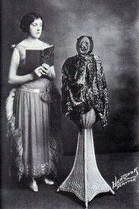 Elsie posing in front of a 1926 microphone, holding a book of fairy tales. Her hair is in tight waves against her head and she is wearing a drop-waist 1920 flapper dress.